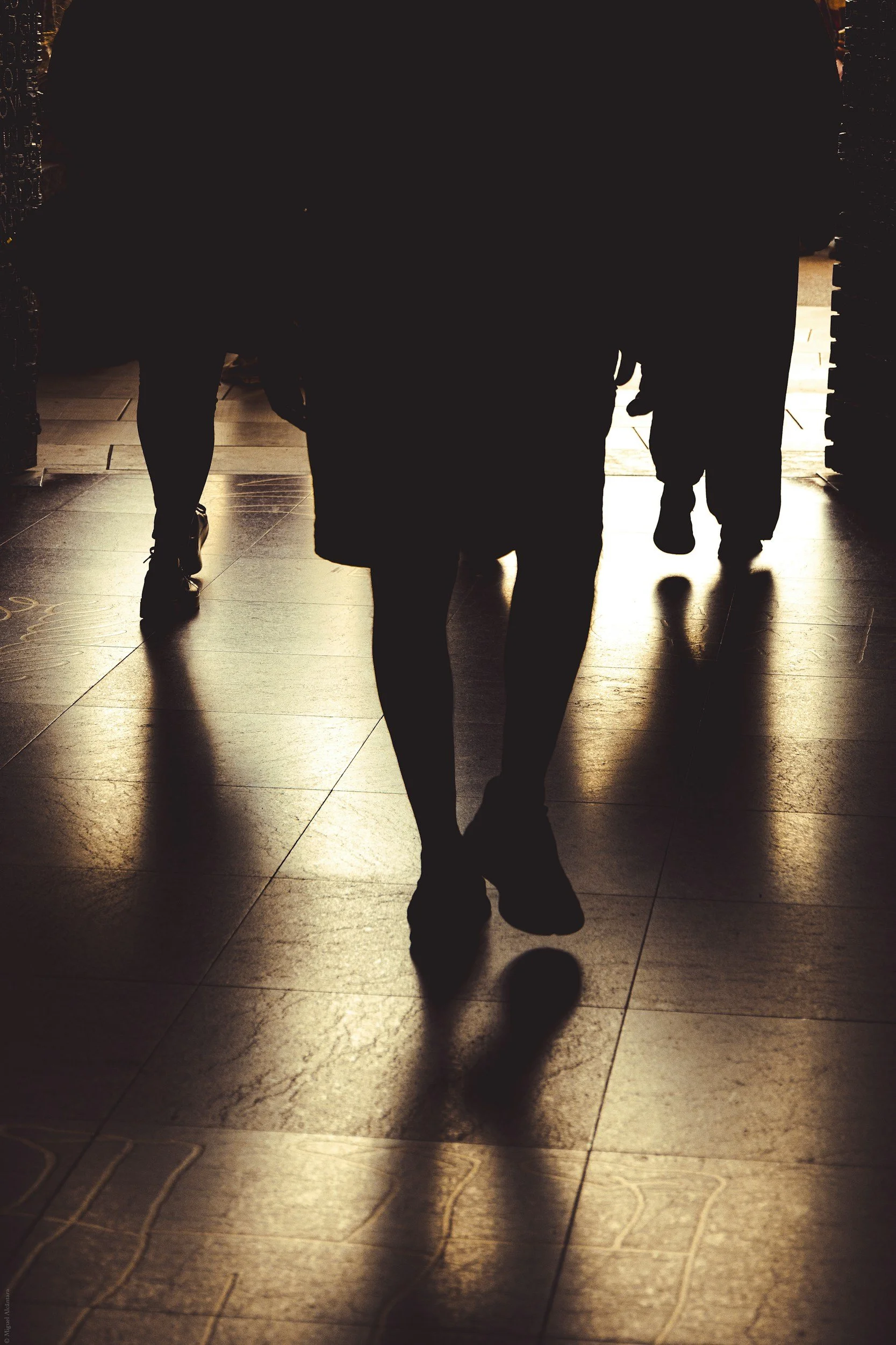 Silhouettes of people walking through a dimly lit doorway, casting long shadows on a tiled floor.