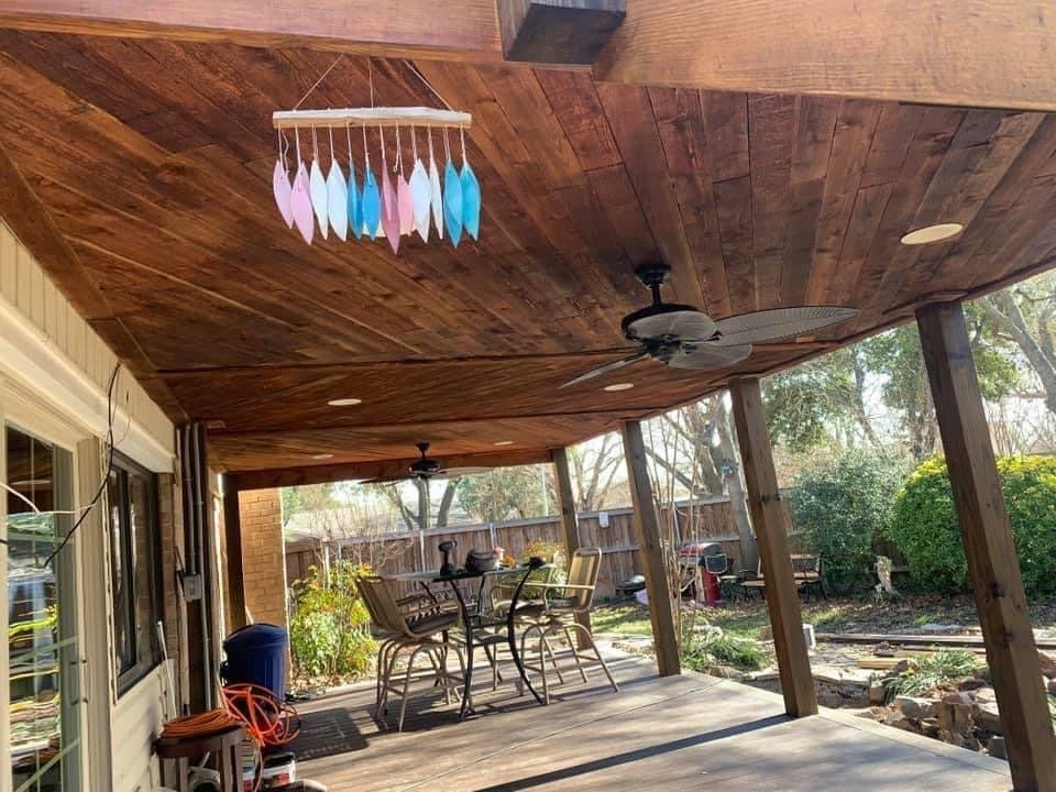 Covered backyard patio with wooden ceiling, ceiling fans, outdoor dining table with four chairs, a colorful feather mobile hanging from the ceiling, and a garden area with shrubs and trees in the background.