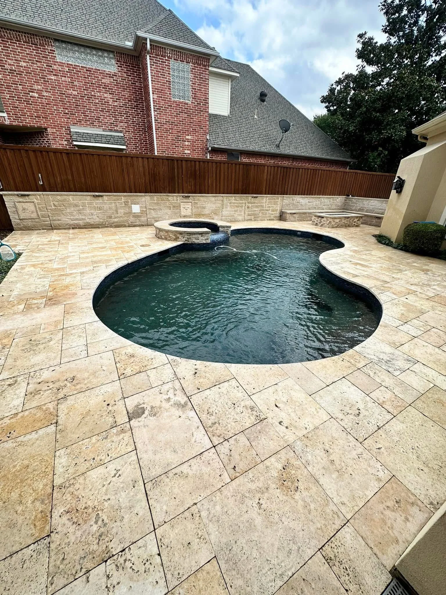 Backyard with a swimming pool, stone patio, wooden fence, brick house, and a wooden bench.