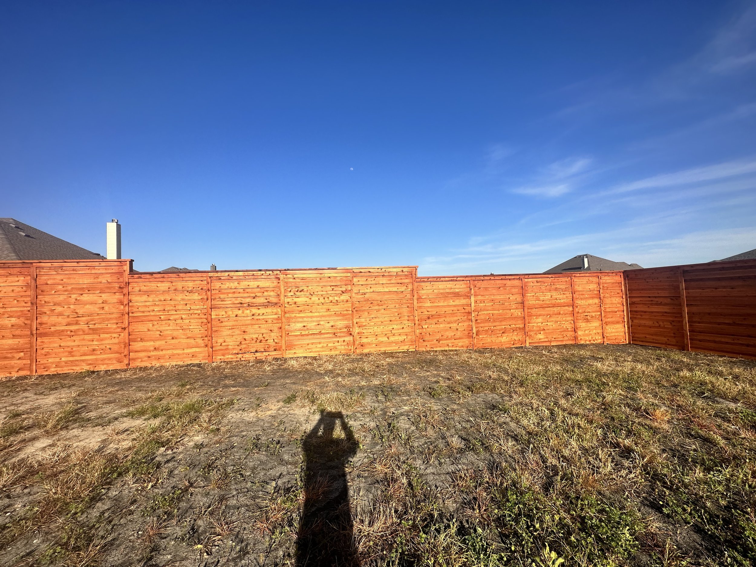 A backyard with a wooden fence, some houses with chimneys, a clear blue sky with the moon visible, and patches of grass and dirt on the ground.