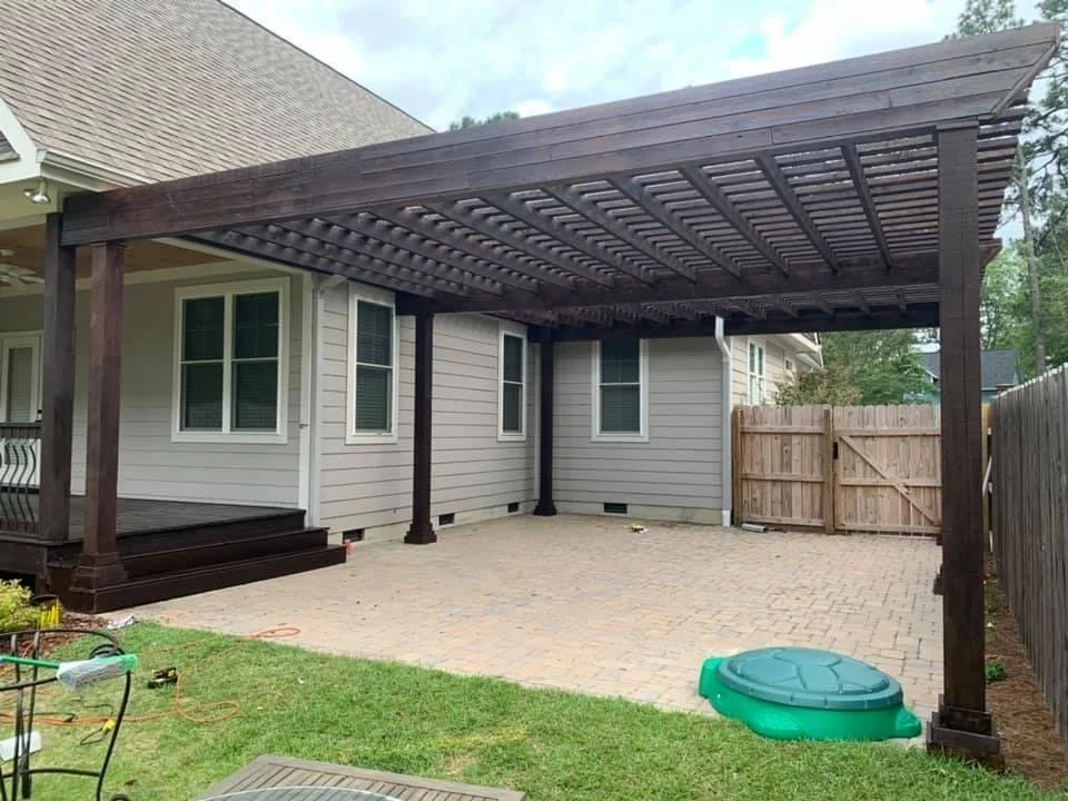 A newly built dark-stained wooden carport attached to a gray house with white trim. The carport is on a brick-paved area with small steps leading up to the house. There is a small fenced backyard with a wooden gate, and a green plastic sandbox on the
