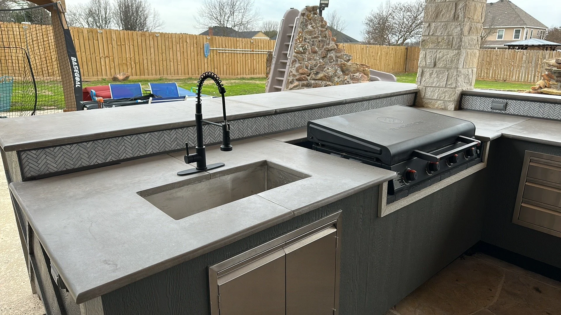 Outdoor kitchen with gray countertops, a black grill, a sink with a black faucet, and a stone barbecue structure in a backyard with a wooden fence and a slide in the background.