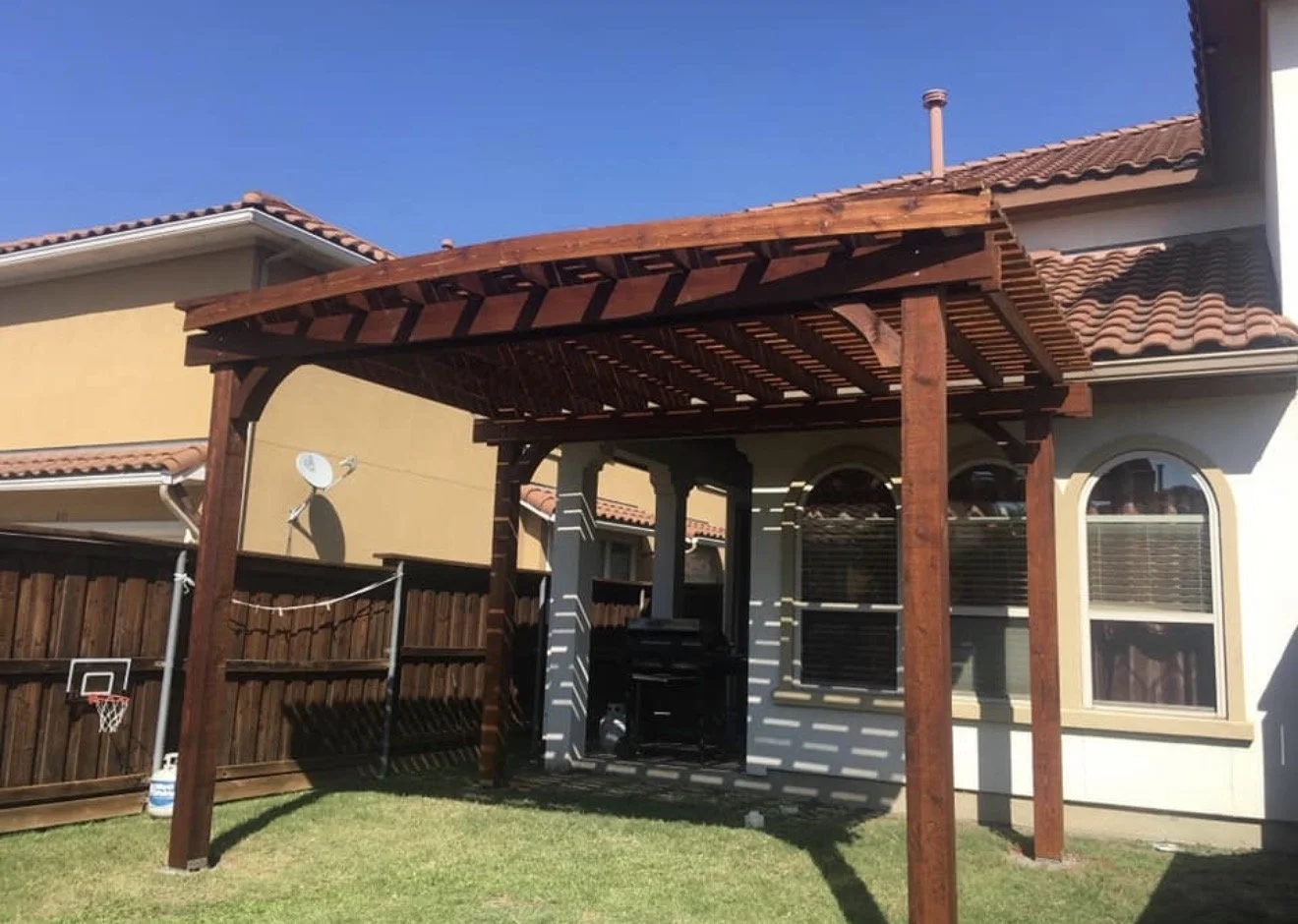 Backyard with a wooden pergola, grass, a small basketball hoop, and houses with tiled roofs in the background.
