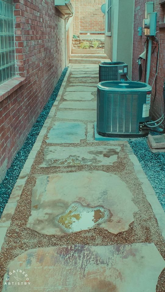A narrow backyard walkway with large stone pavers, bordered by gravel, running between a brick house and utility equipment, leading toward steps at the back.