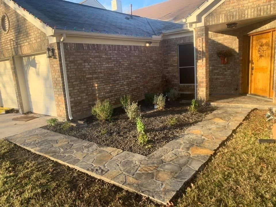 Front yard with a stone pathway surrounding a small garden bed with plants, brick house with a wooden door, and garage with a white door.