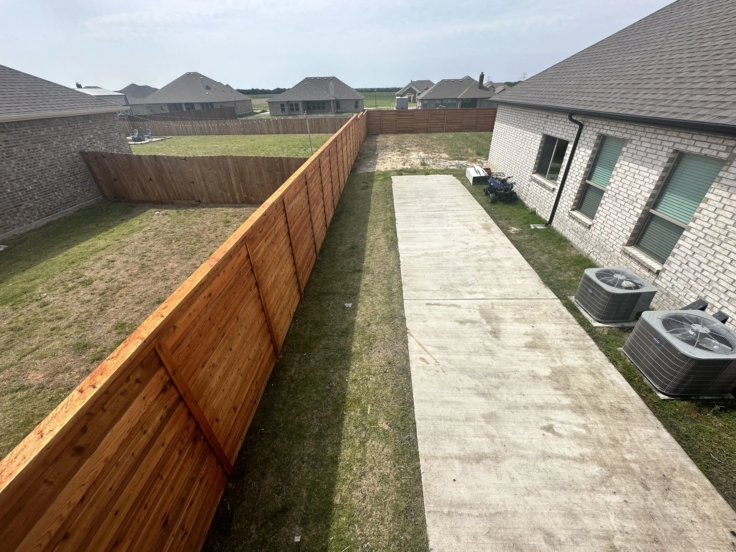 A backyard with a wooden fence, a concrete pathway, and some lawn space, with neighboring houses in the background.