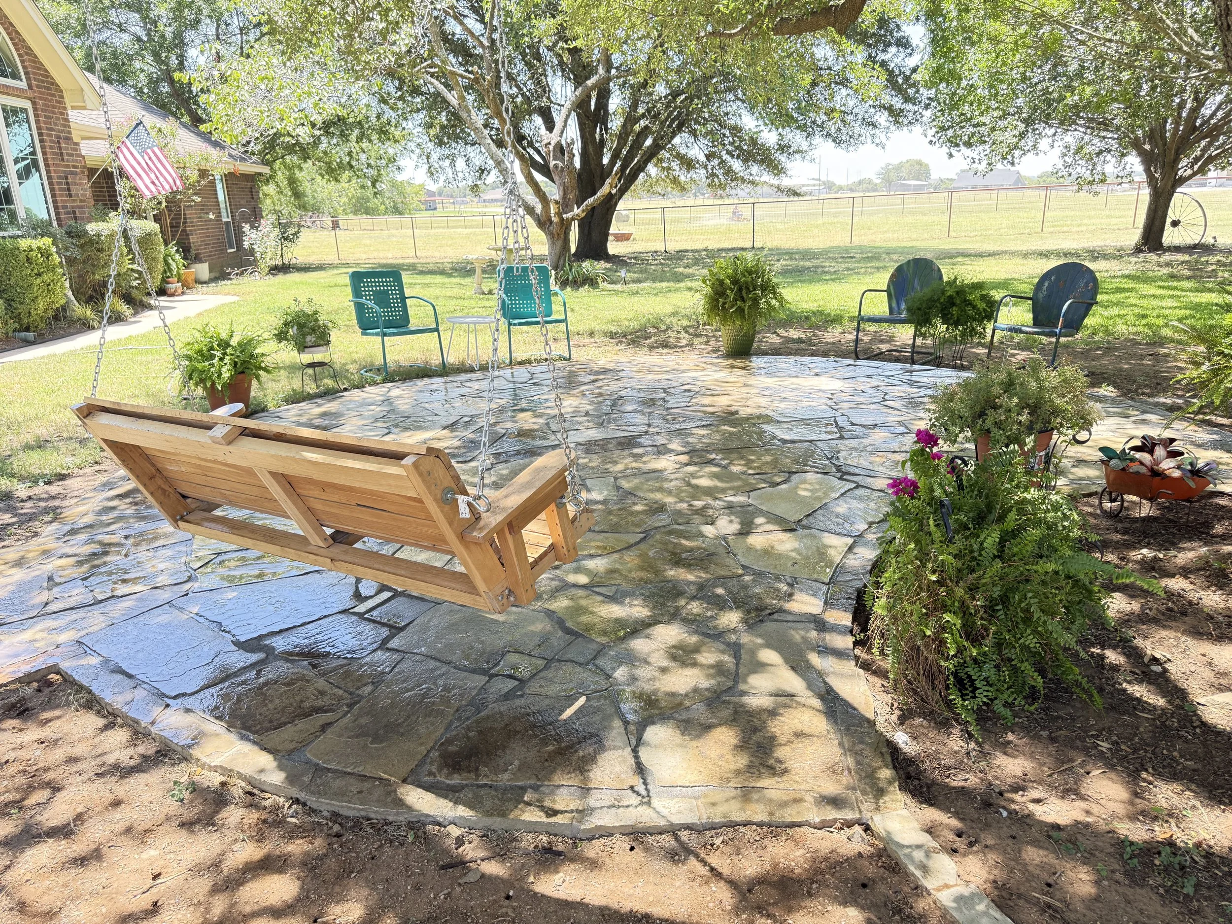 Backyard with a wooden swing, two chairs, potted plants, large trees, and a stone patio with a grassy field in the background.