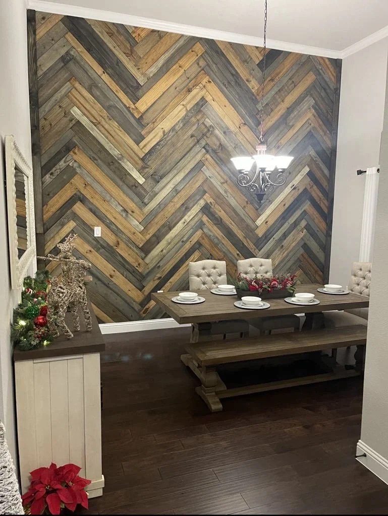 A dining room with a rustic chevron wood accent wall, a wooden dining table with six beige upholstered chairs, a chandelier hanging above, and holiday decorations including a red poinsettia and Christmas ornaments.