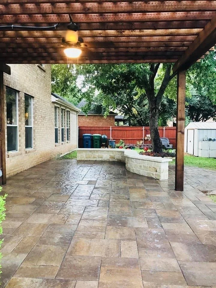 A covered patio with brown tiled flooring, a brick house wall with multiple windows, a ceiling fan with a light, and a garden area with a tree, plants, and a red wooden fence in the background.