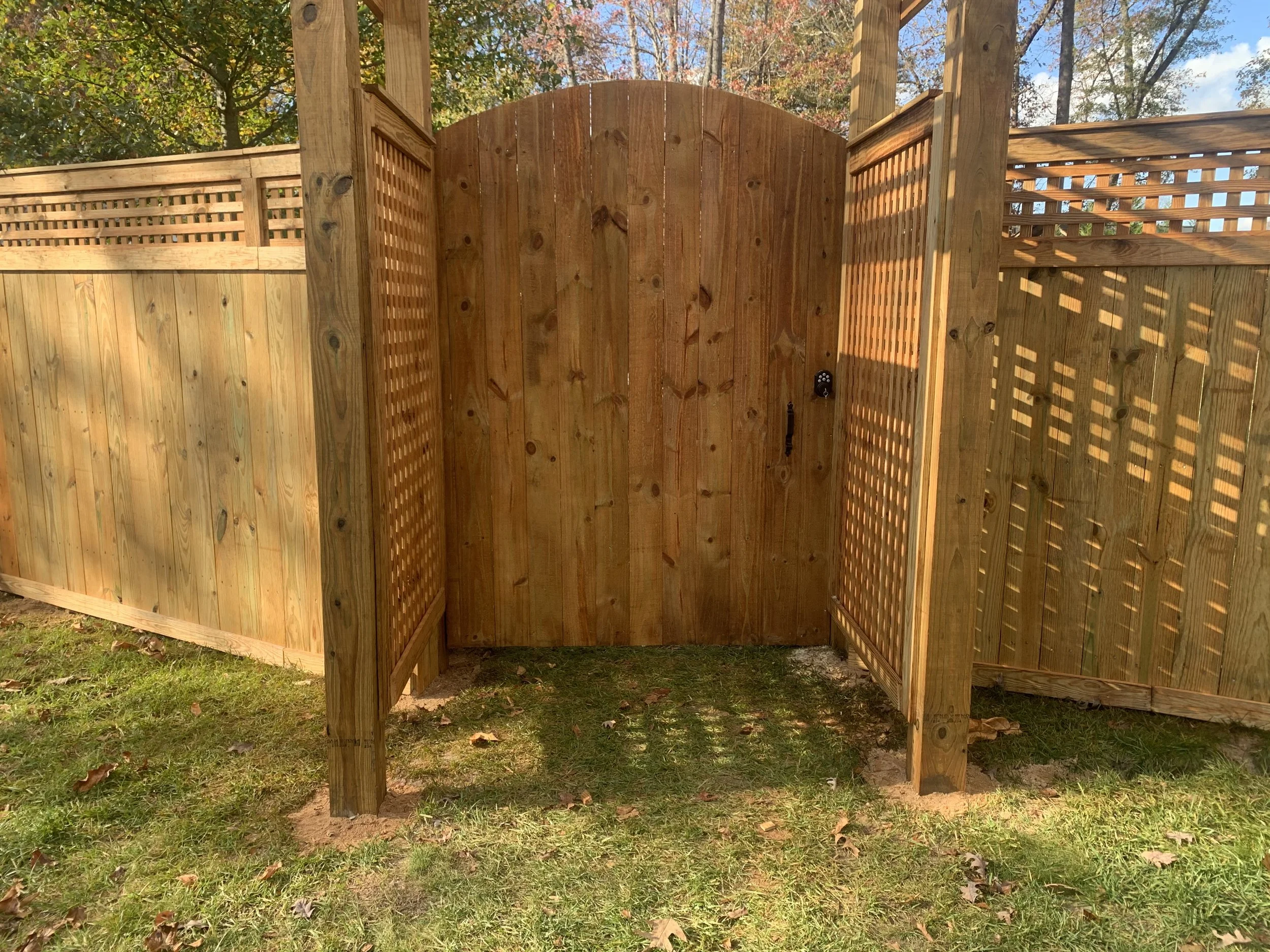 Wooden outdoor shower with a closed door and lattice panels on each side, set in a yard with grass and trees in the background.