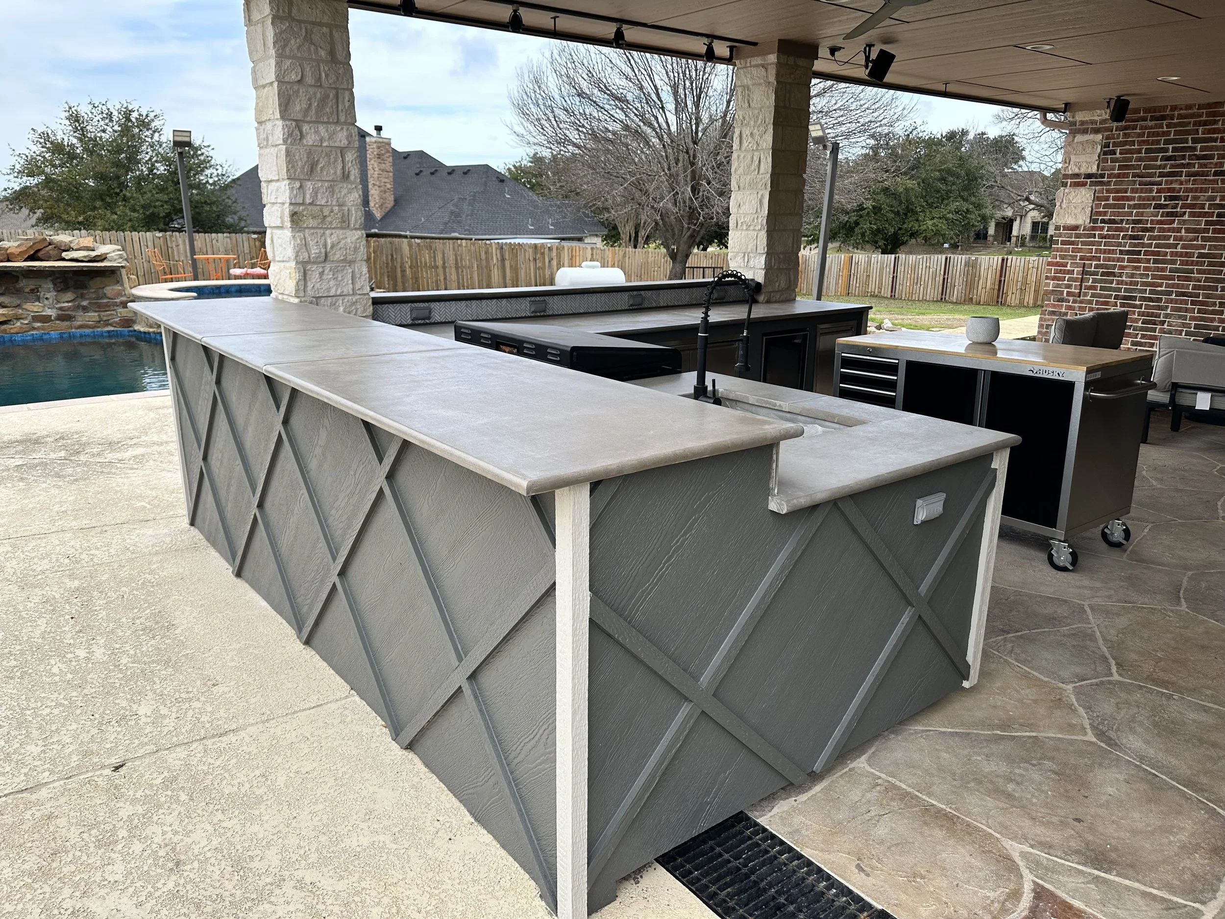 Outdoor kitchen with a stone bar, black sink with a pull-down faucet, grill, and a dessert cart with a wooden top, set under a covered patio with stone columns, brick wall, and a backyard with a fence, trees, and pool in the background.