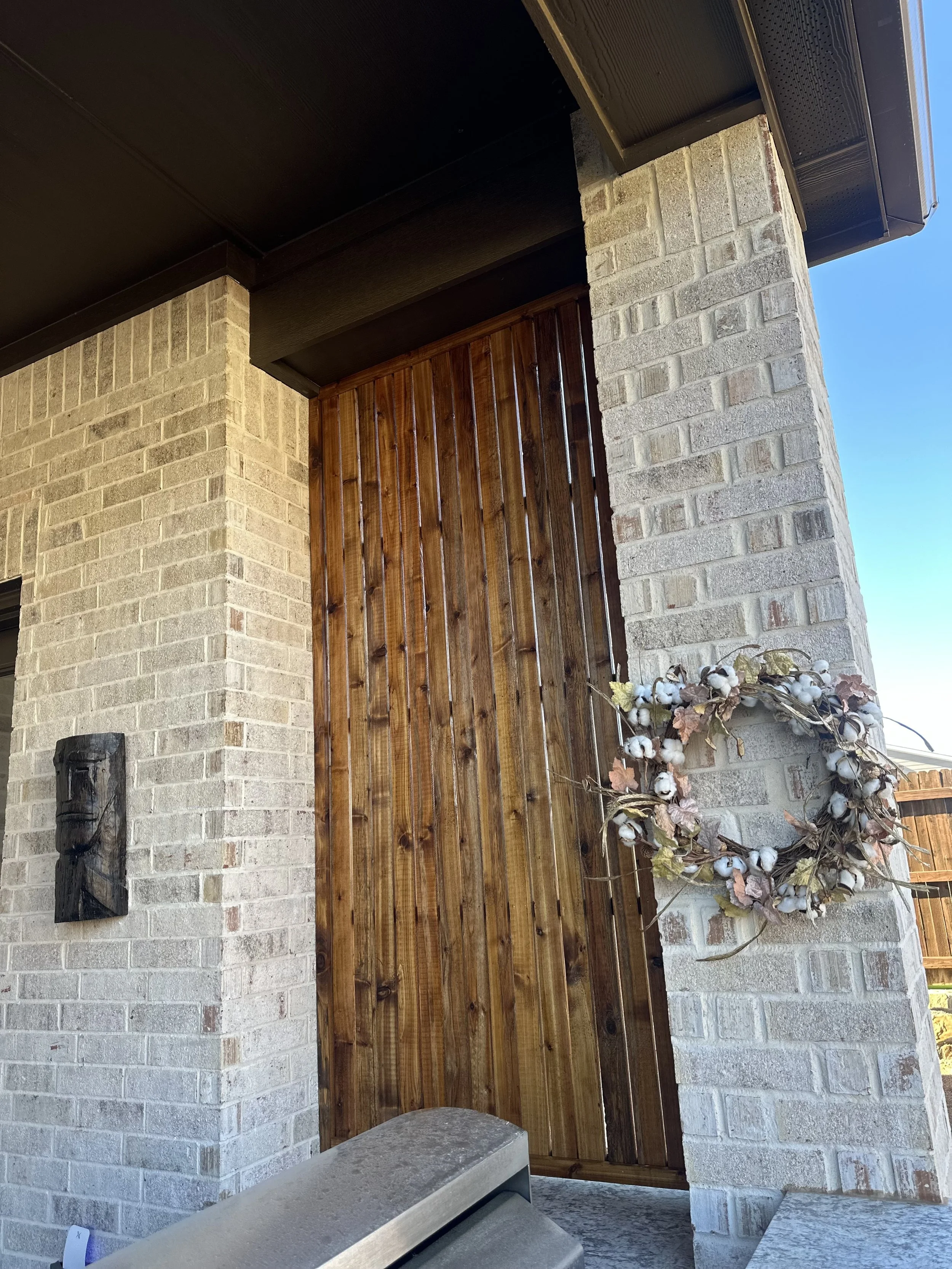Exterior view of a house showing brick walls, a wooden door, and a decorative wreath made of cotton stems and dried leaves hanging on the wall near the door.