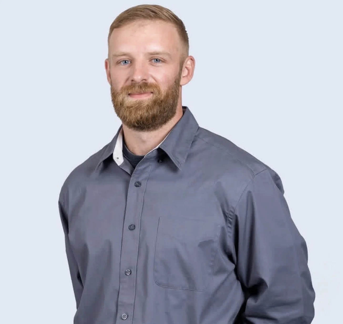 A man with light skin, a red beard, and short red hair, wearing a gray button-up shirt, standing against a light gray background.