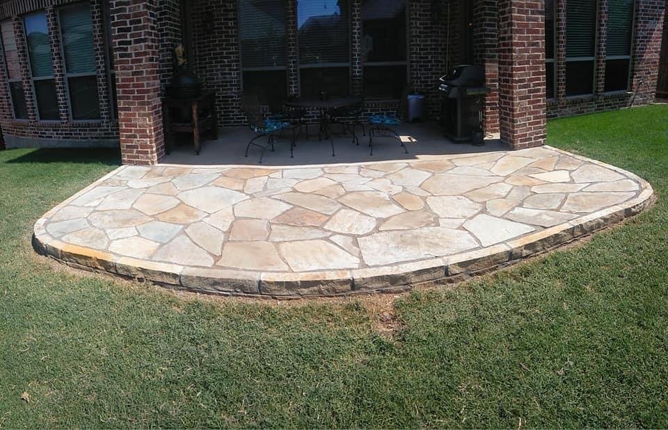 A stone patio with a curved edge, attached to a brick house with a covered back porch, outdoor furniture, and a BBQ grill.