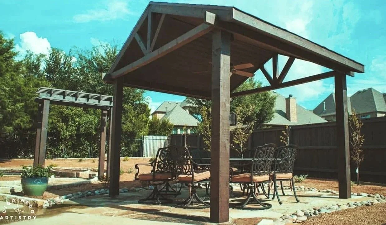 Backyard patio with a wooden pergola, metal outdoor furniture, and a small garden area with trees and a fence.