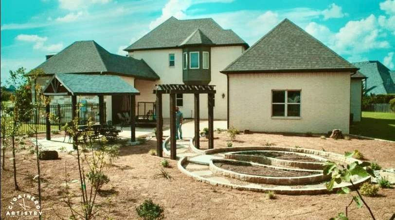 A backyard with a new house in the background, a small covered patio area with outdoor furniture, a spiral-shaped garden bed, and newly planted young trees and shrubs.