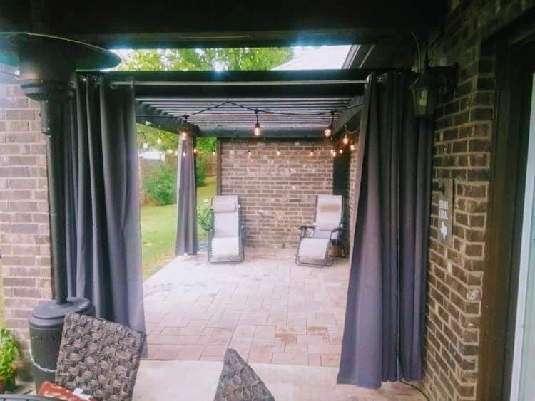View of a covered outdoor patio with brick walls, two lounge chairs, string lights, and a table with chairs, seen through an open sliding door with dark curtains.