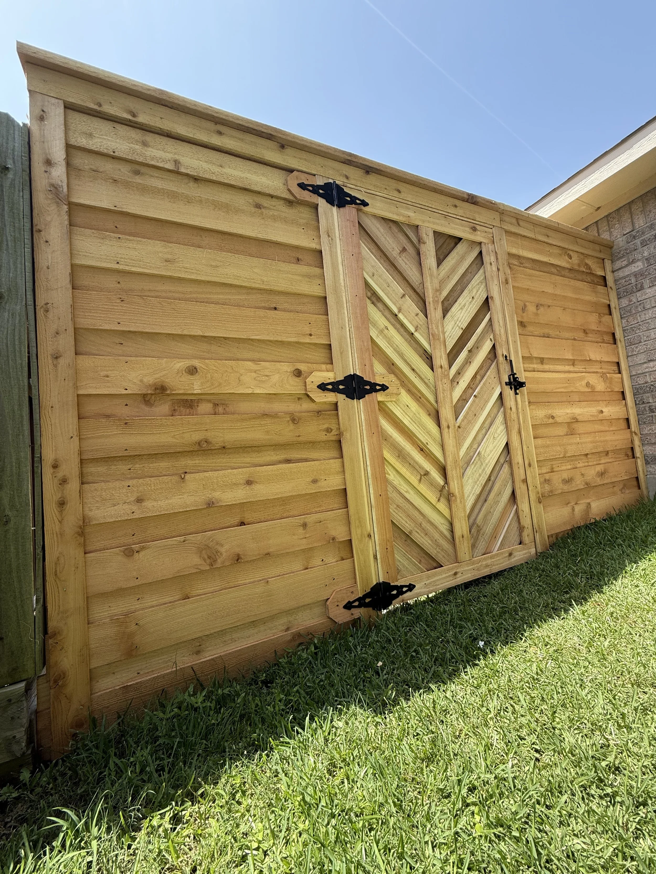 A wooden shed with a boarded door and black decorative hinges, situated on a grassy lawn with a brick house on the right and a blue sky above.
