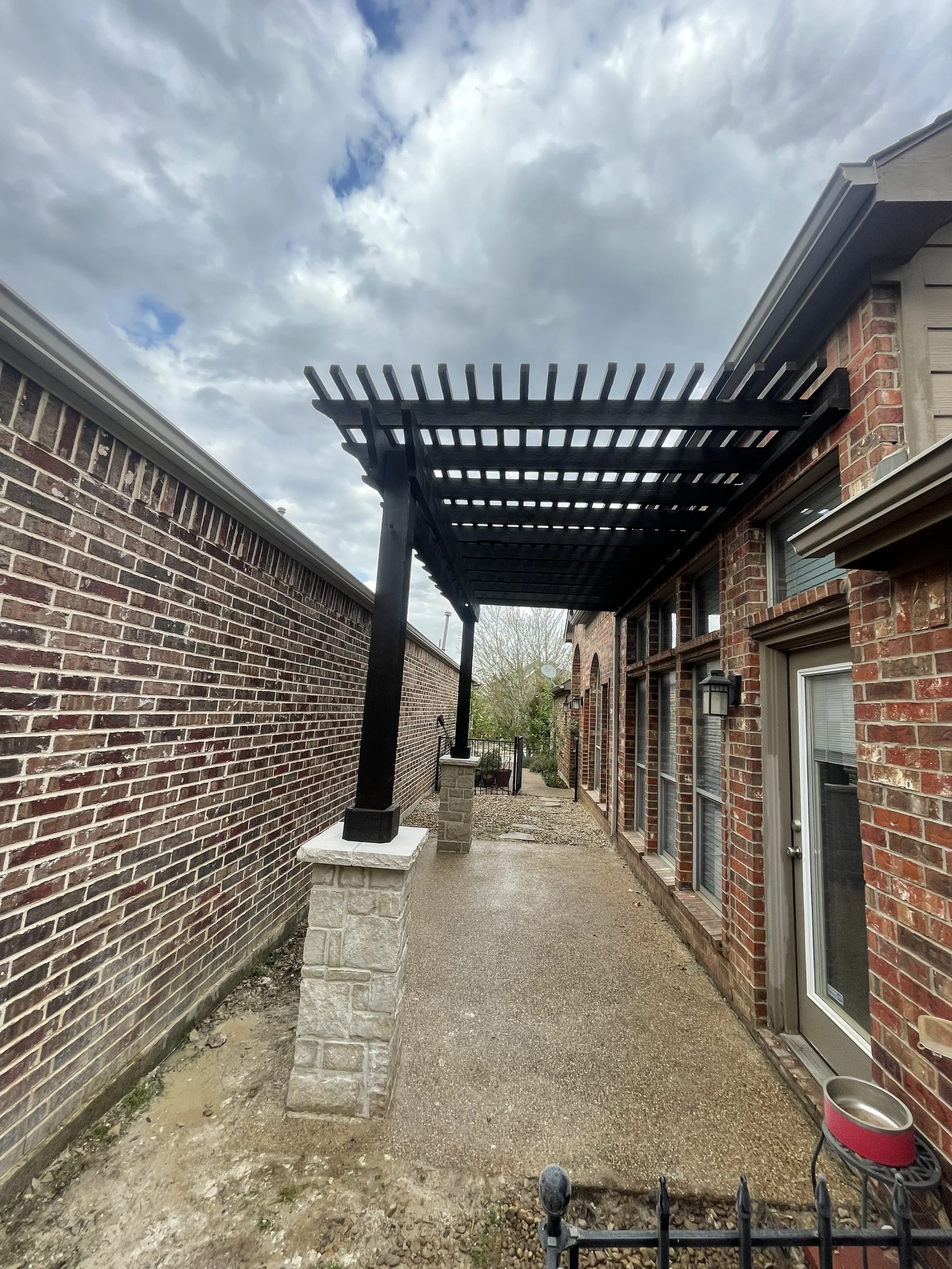 Backyard patio area with a brick wall on the left and a brick house on the right side. In the center, there's a black pergola partially extending from the house, supported by columns with stone bases. The ground is a mix of dirt and gravel. The sky i