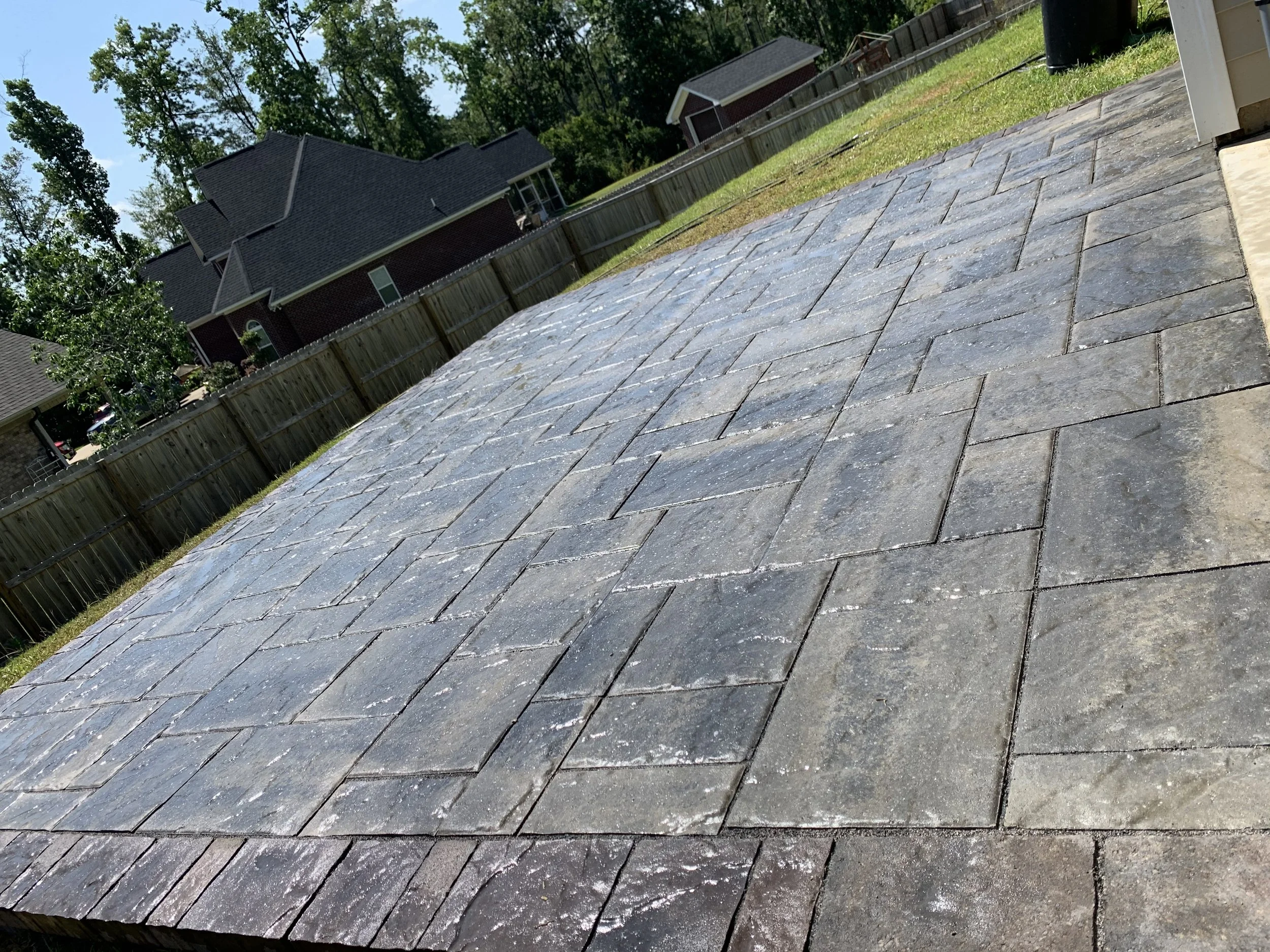 A backyard patio with gray stone pavers, surrounded by a wooden fence, and a house with dark roofing and brick siding in the background. There are trees and a grassy yard beyond the fence.