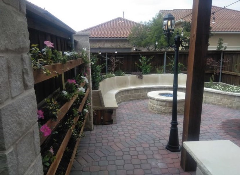 Backyard patio with brick pavers, a curved stone bench, a fire pit, a black lamppost, string lights, and a wooden fence with flower pots.