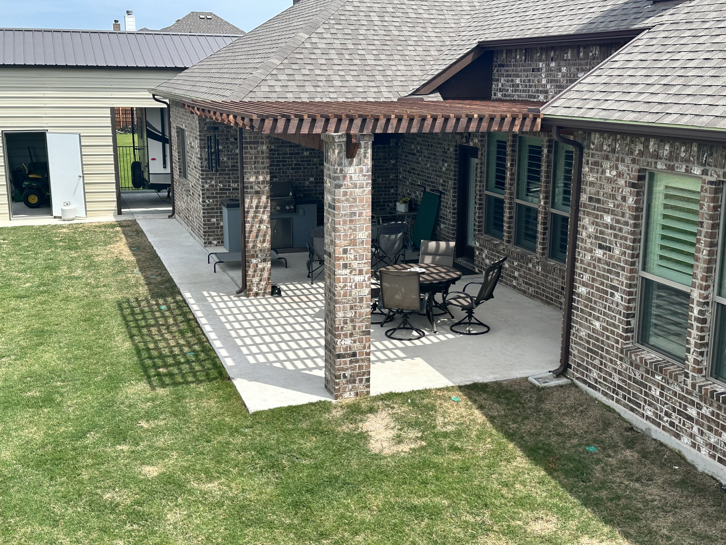 A backyard patio area with a brick house, concrete floor, and outdoor furniture under a wooden pergola. A green lawn surrounds the patio.
