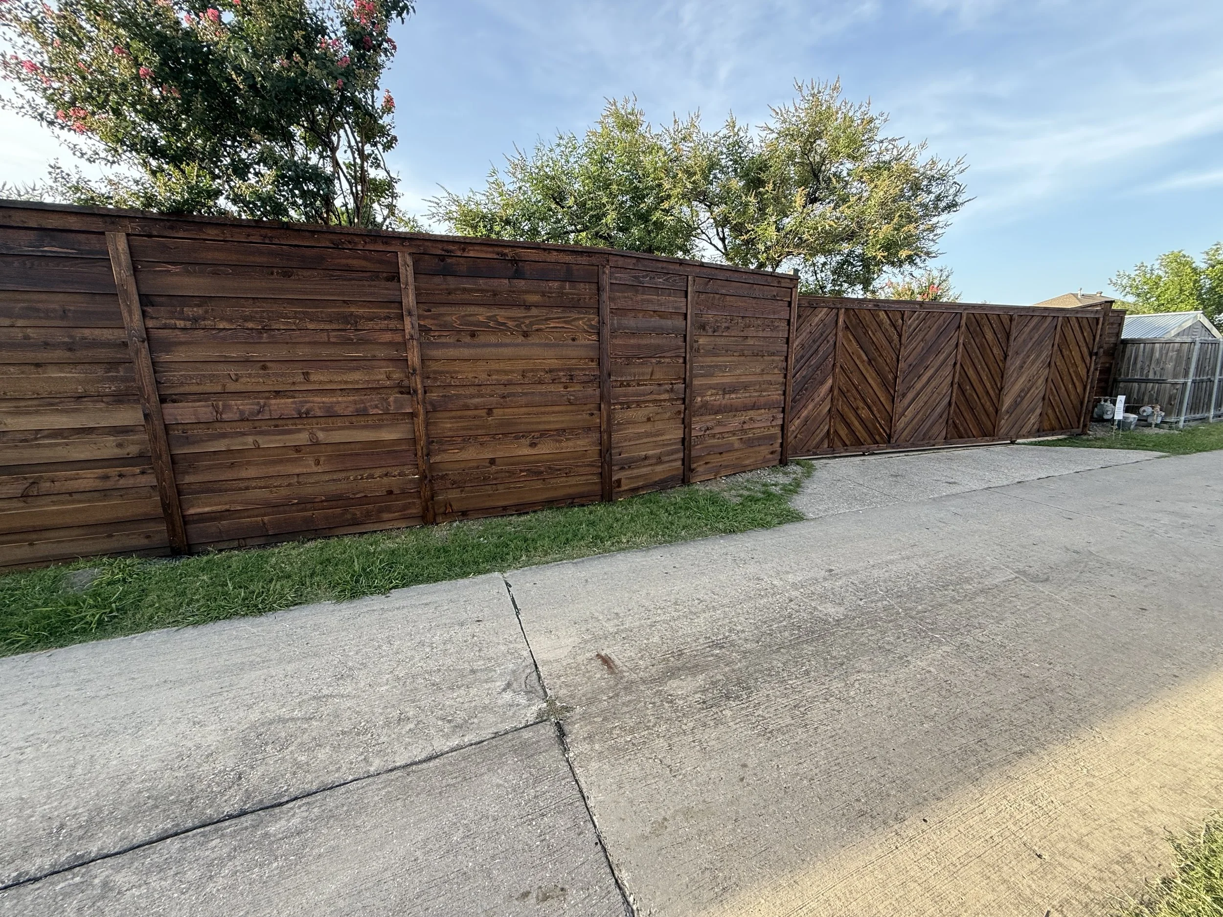 A wooden privacy fence along a concrete sidewalk. In the background, there are trees with green leaves and a house with a metal roof. The sky is partly cloudy and blue.