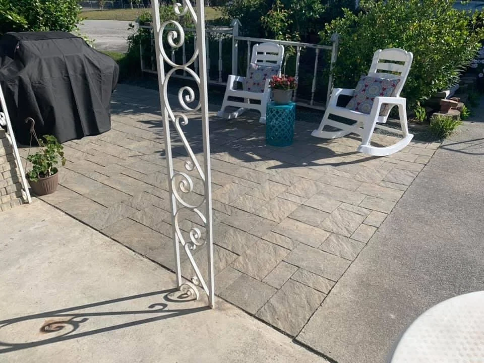 A small outdoor patio area with a stone-tiled surface. There are two white rocking chairs with pillows, a small blue side table, and a potted plant. A decorative white metal post is in the foreground, casting a shadow. The background has greenery and