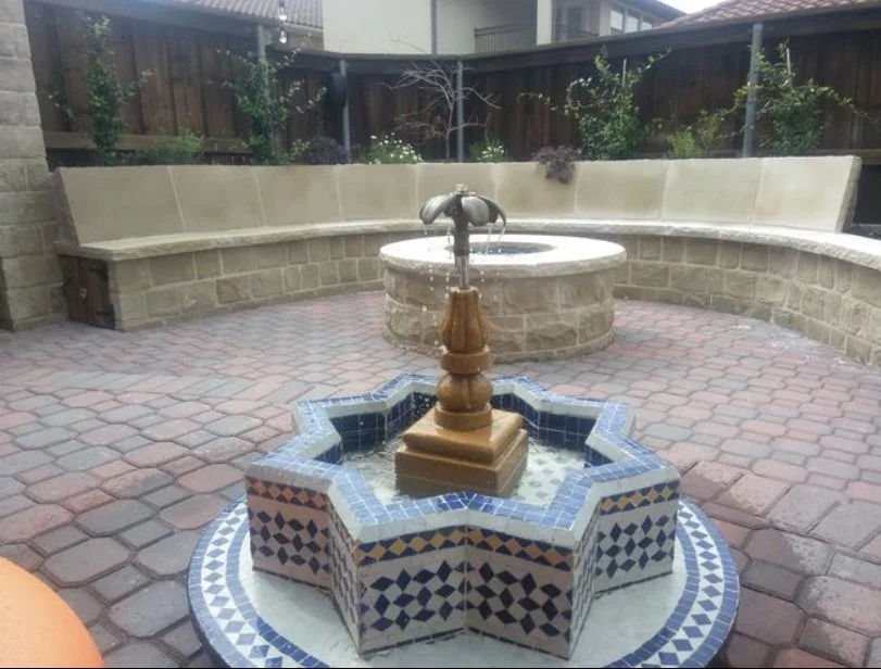 A backyard patio with a two-tiered stone fountain with a tiled, star-shaped base, and a larger circular stone fountain in the background, surrounded by a stone bench and decorative plants.