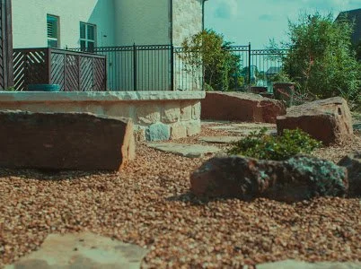 A yard with a stone and gravel landscape, surrounded by a metal fence, with plants and a house in the background.