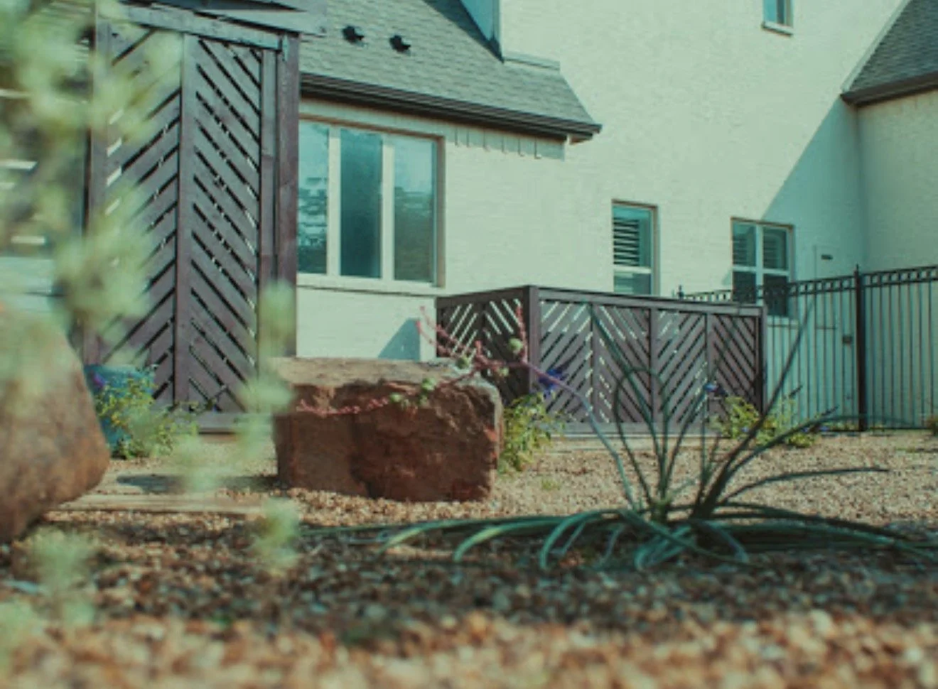 View of a backyard with gravel ground, a small plant, a brick, and a wooden deck with a railing in front of a house with white walls, multiple windows, and a sloped roof.