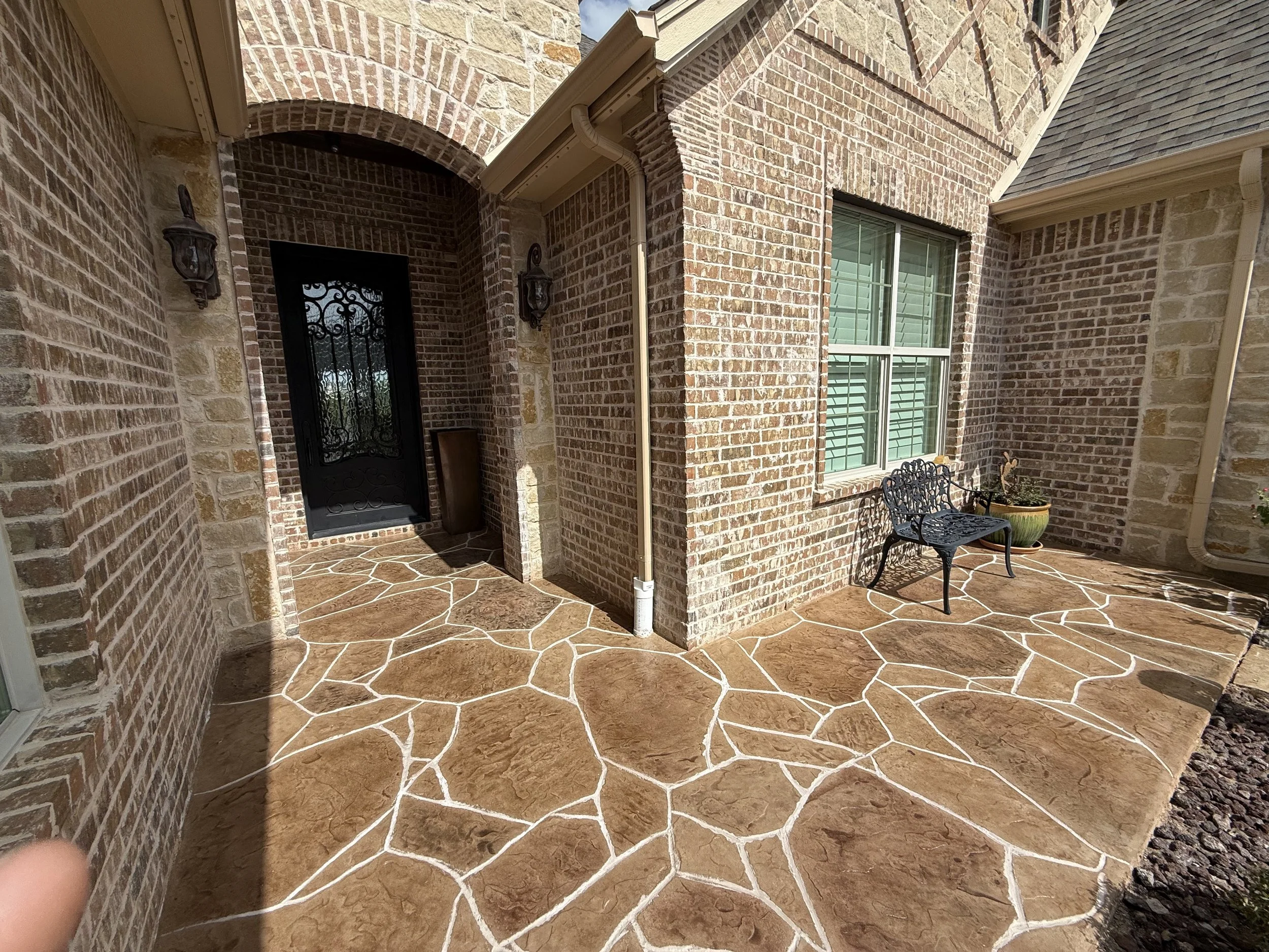 Exterior view of a brick house porch with a black metal door and window, decorated with a black outdoor lamp and potted plants, featuring a high end concrete overlay to look like real flagstone.
