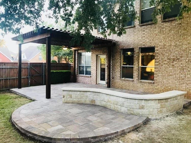 A backyard patio with a curved stone wall, a paved surface, a small covered area, and a brick house with multiple windows.