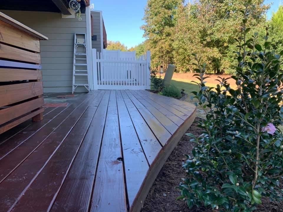 A wooden deck attached to a house, with a white picket fence in the background and a garden bed with green bushes on the right. The deck is wet and appears newly stained, with sunlight and clear skies visible.