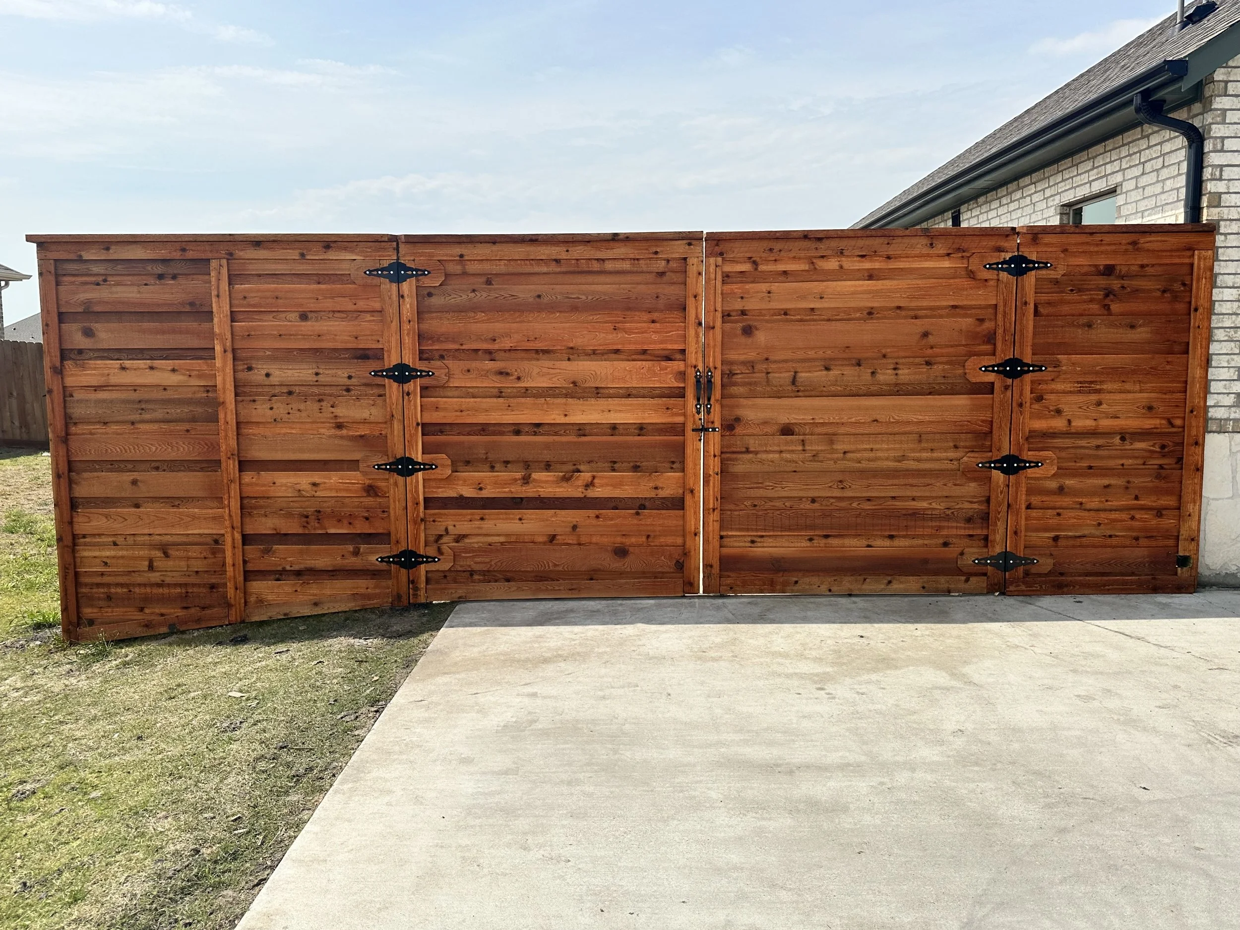 New wooden privacy fence with black hardware installed along a house with brick exterior, overlooking a concrete patio and grassy yard under a partially cloudy sky.