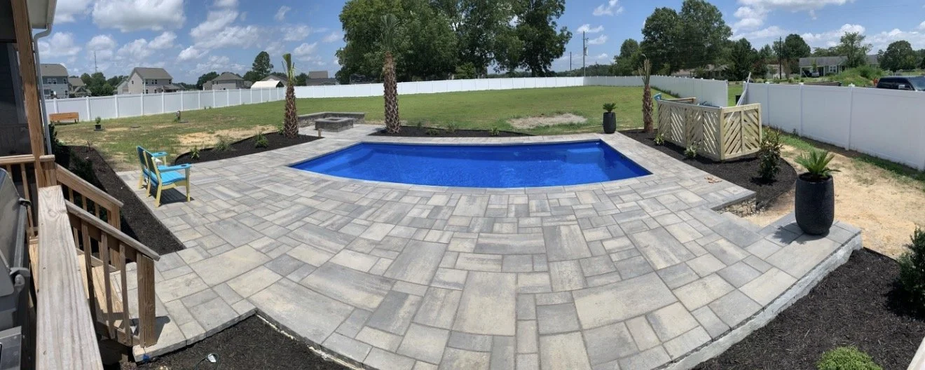 Backyard with a swimming pool, gray brick patio, two palm trees, two blue chairs, black planters with greenery, white fence, grassy area, and a partly cloudy sky.