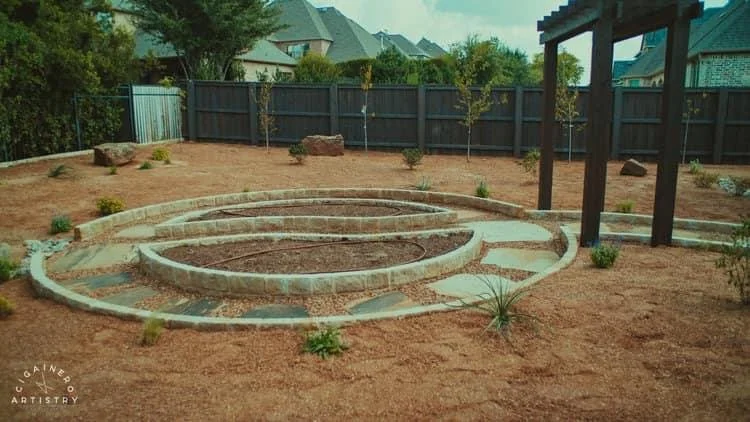 A backyard with a circular staircase made of concrete and brick, and a gravel pathway leading to a small wooden pergola, surrounded by brown mulch and small plants, enclosed by a black fence.