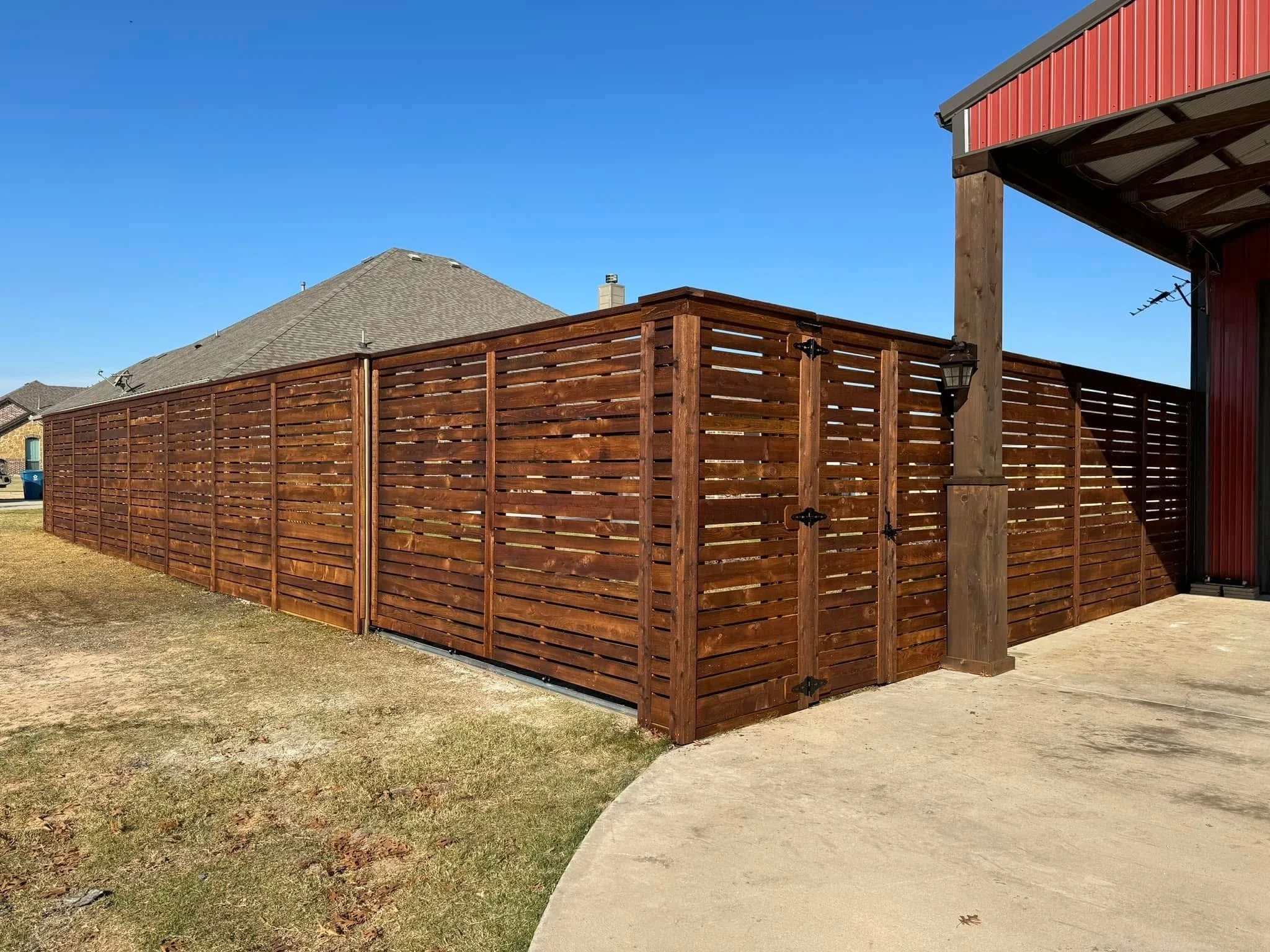 A backyard with a wooden privacy fence, a concrete patio, and a covered area. The roof of the house is visible in the background with clear blue sky overhead.