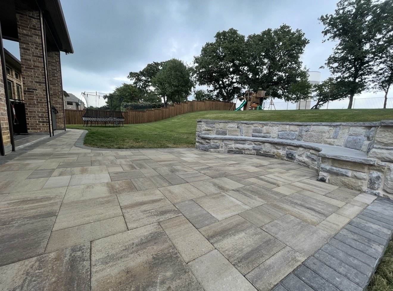 Backyard patio with stone flooring, a stone retaining wall, a grassy lawn, trees, a wooden fence, and a children's playset with a slide in the background.