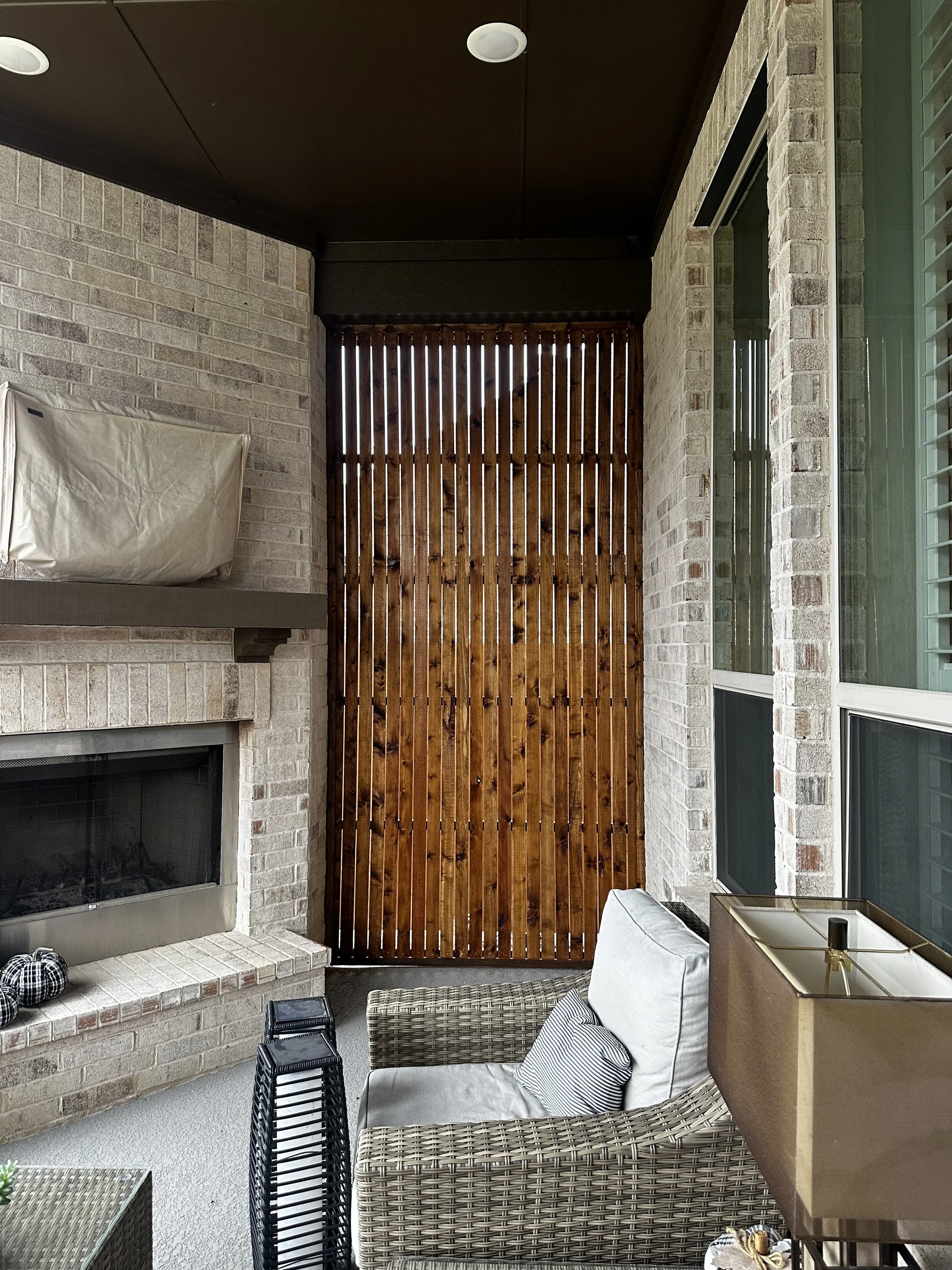 A cozy outdoor patio area with wicker furniture, including a loveseat with cushions, a small table, and stacking chairs. There is a brick fireplace with a mantel on the left, a white pillow on the loveseat, and a decorative box on a side table. The a