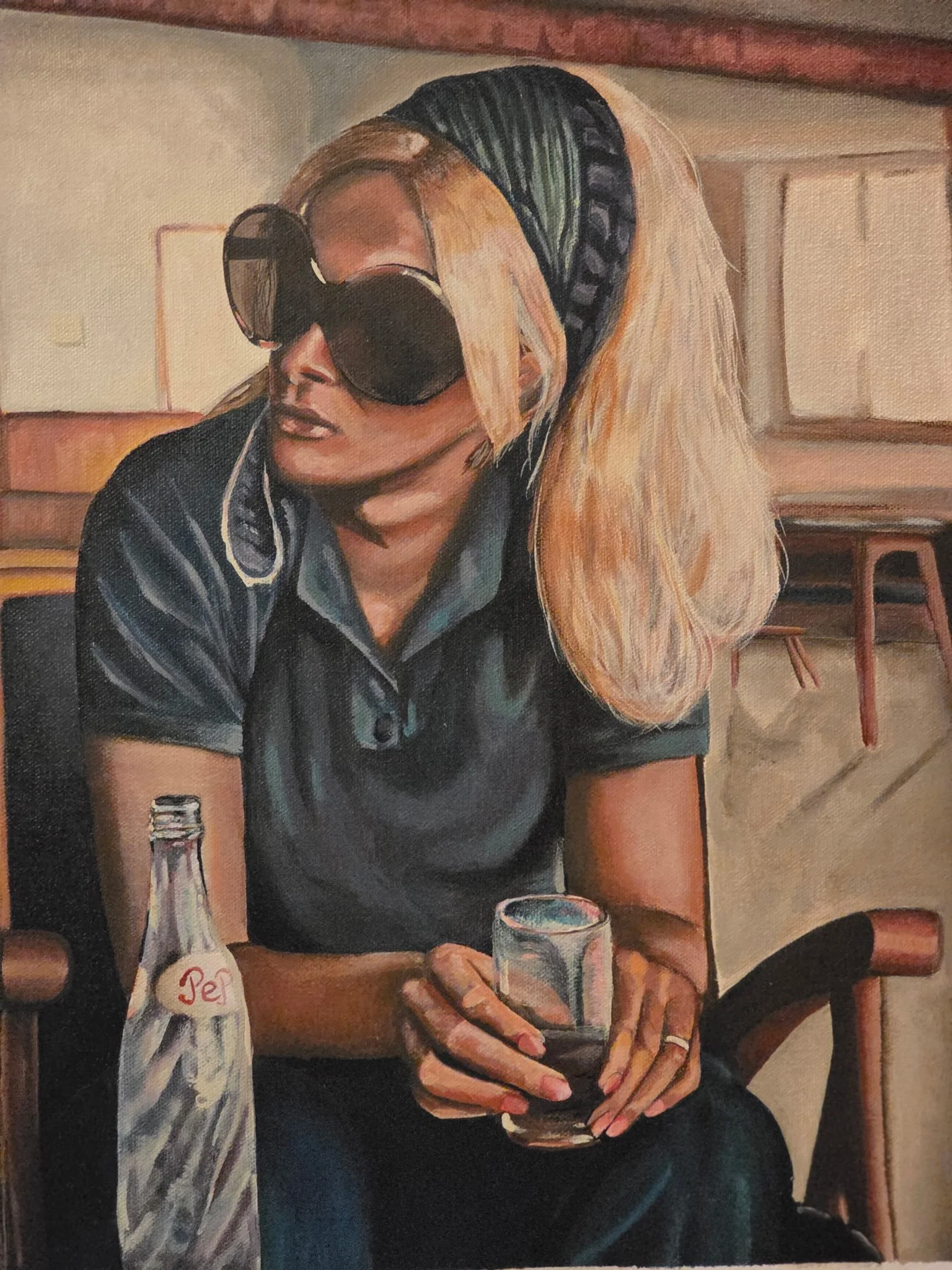 A woman with long blonde hair wearing large sunglasses, a striped top, and a black cardigan sitting at a wooden table in a cozy room. She is holding a glass of dark beverage and a pack of Pez candies is on the table.