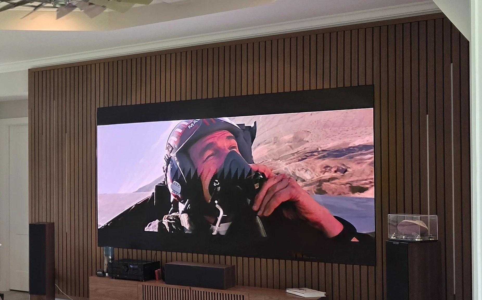 A large flat-screen TV mounted on a wall with wooden paneling, displaying a close-up of a pilot in a helicopter wearing a helmet, with a desert landscape in the background.