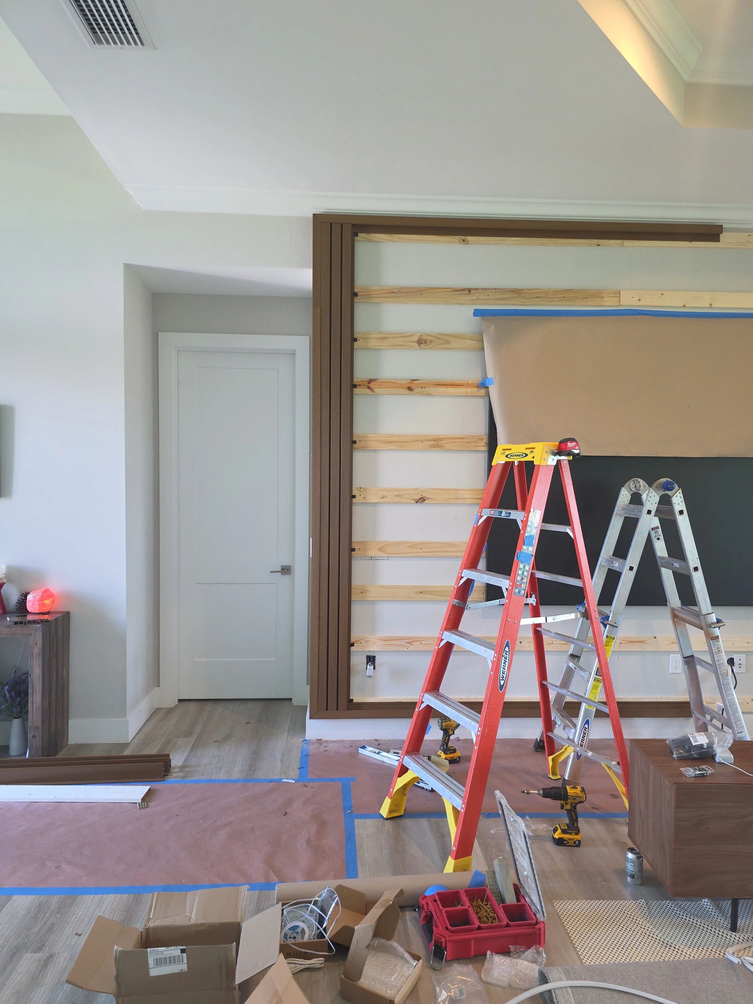 Interior of a room undergoing renovation with ladders, tools, and construction materials scattered on the floor, partially built wooden frame on the wall, and a closed white door.