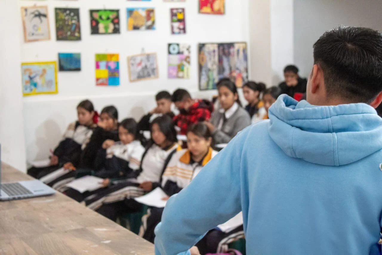 Un instructor frente a un grupo de estudiantes en un aula, con obras de arte en la pared.