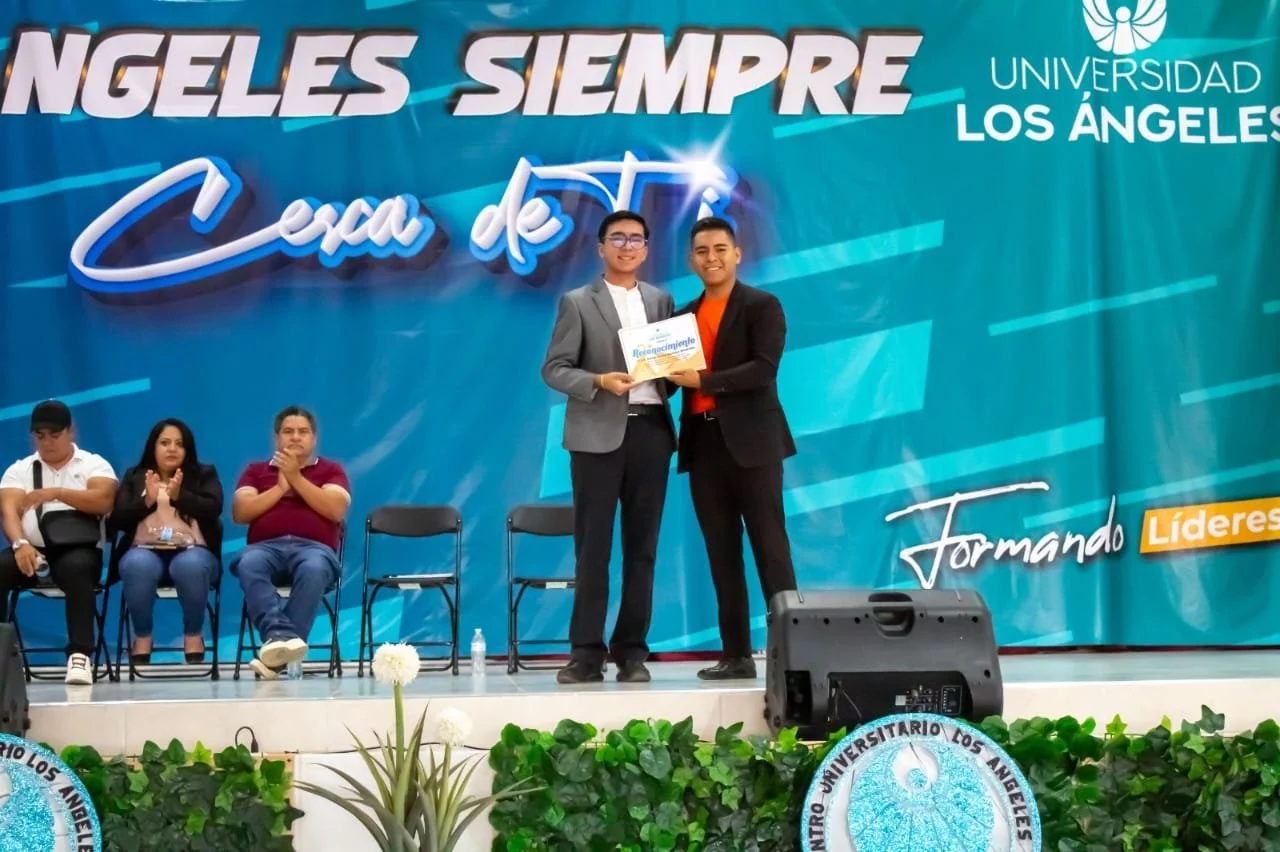 Dos personas en el escenario en una ceremonia de reconocimiento en la Universidad Los Ángeles, con un fondo azul y el texto 'Ninglés Siempre' y 'Cear de ti'. Una de ellas está recibiendo un diploma. Hay otras personas sentadas en las sillas del escenario y plantas verdes decorando el frente.