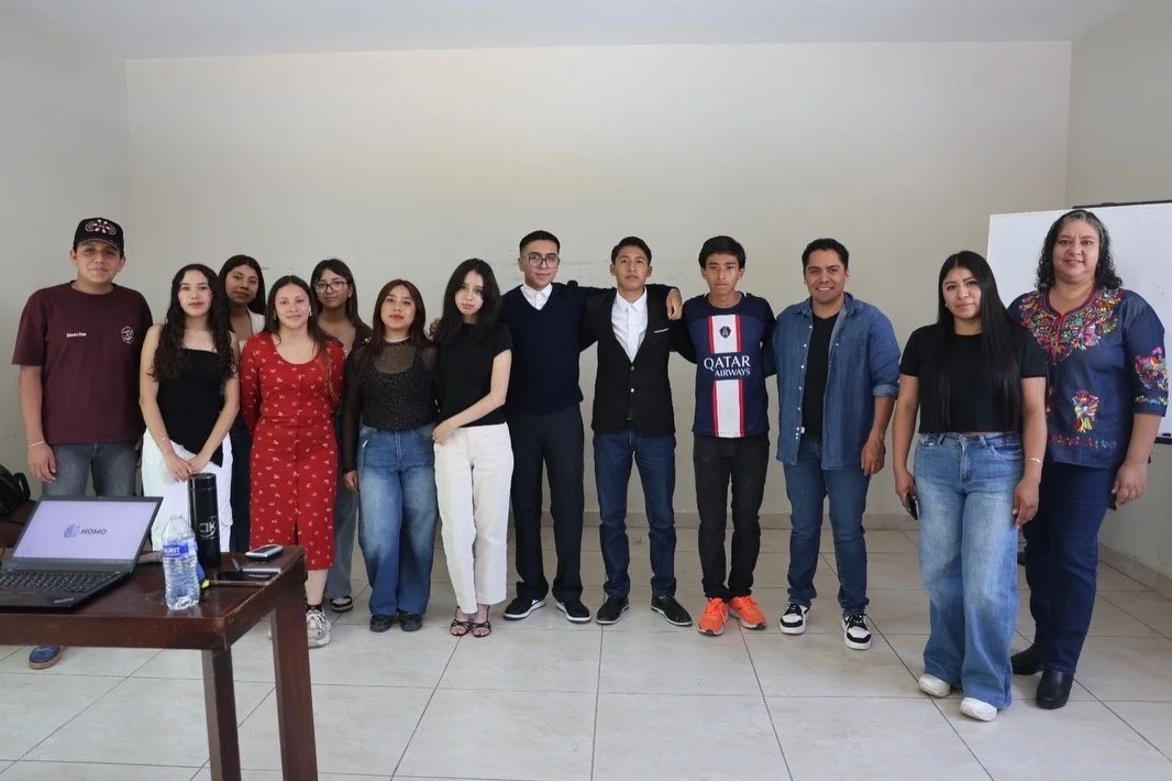 Grupo de estudiantes y una docente posando en un salón de clases con equipo de computo y pizarra.