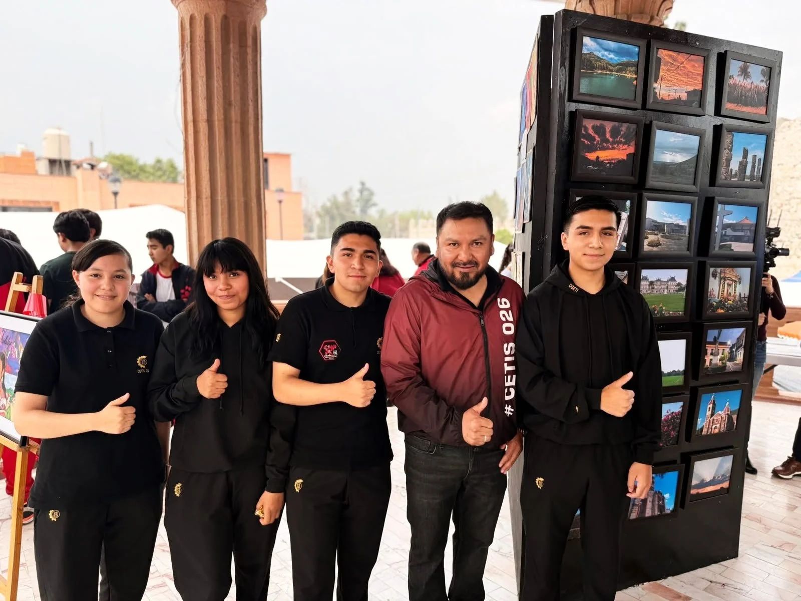 Grupo de cinco jóvenes y un adulto, en una exhibición, posando para la foto y haciendo señal de pulgar arriba frente a una estructura con muchas fotografías de paisajes y lugares icónicos.