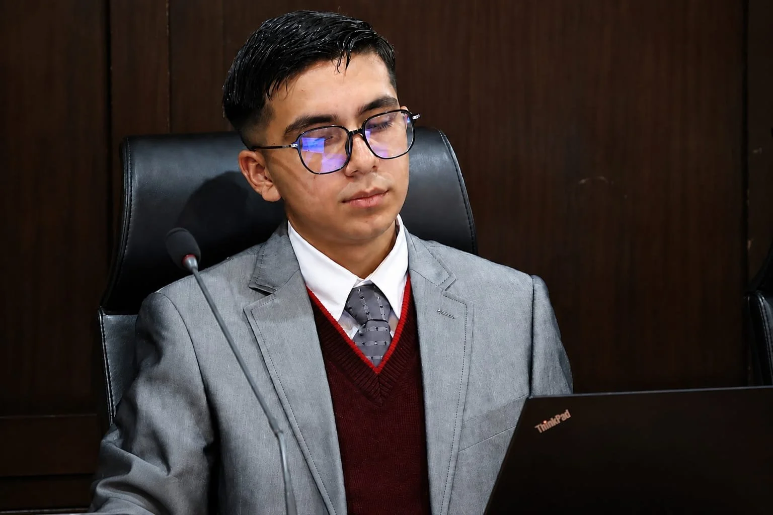 Hombre joven con gafas, vestido con saco gris, corbata y suéter burdeos, sentado en una silla frente a una laptop en un ambiente formal.