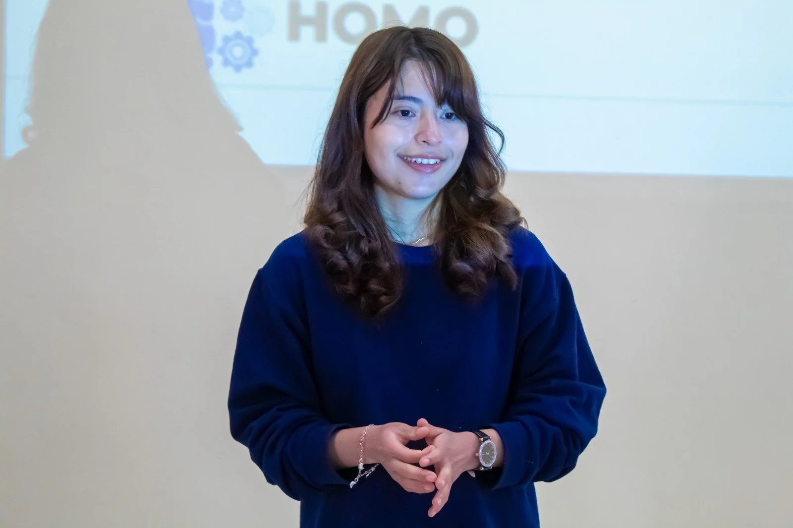 Joven mujer con cabello rizado, camisa azul, sonriendo y en un escenario con una pantalla detrás que tiene la palabra 'HOMO'.