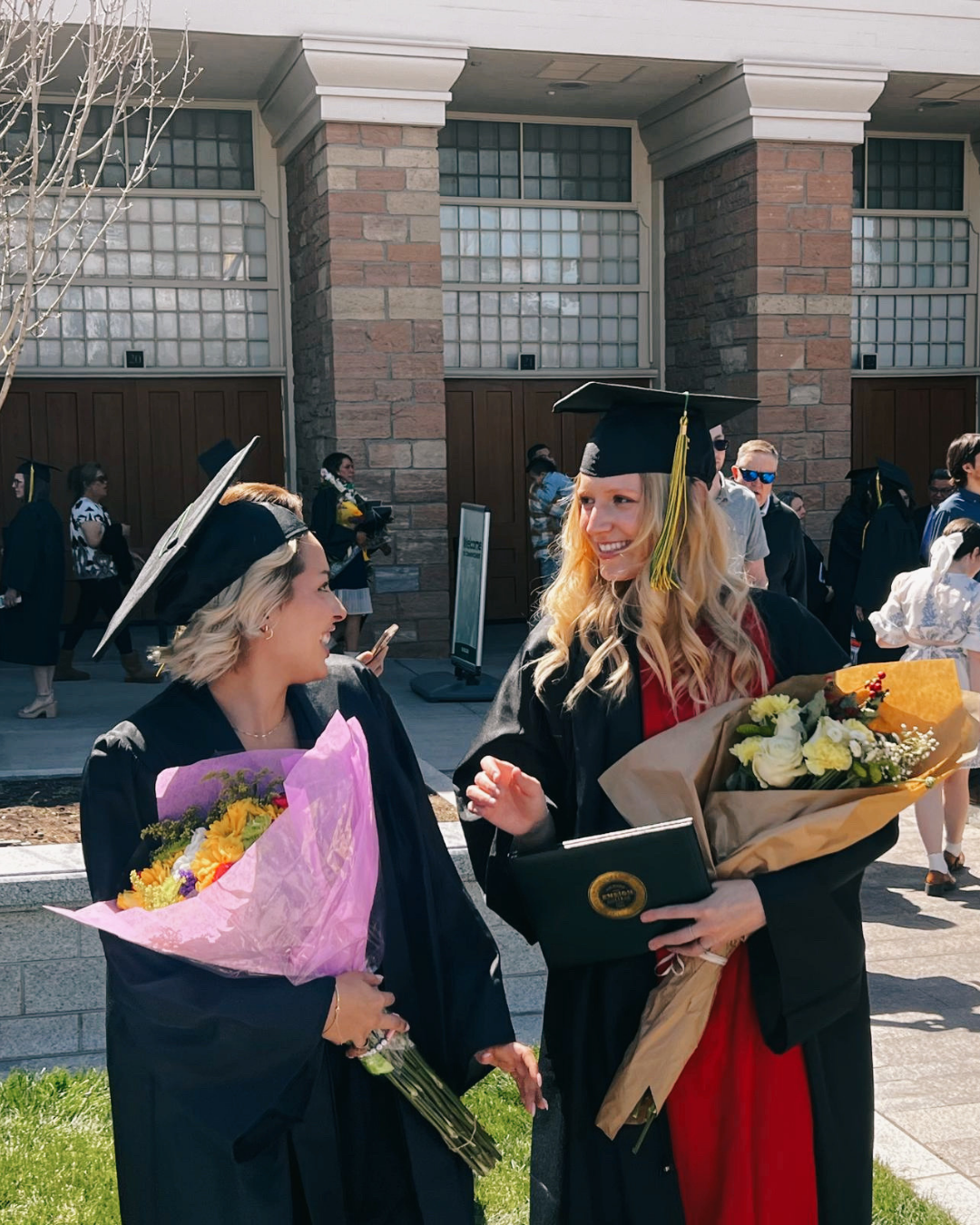 Two women in graduation caps and gowns holding bouquets of flowers talking to each other outside, with other graduates in the background.